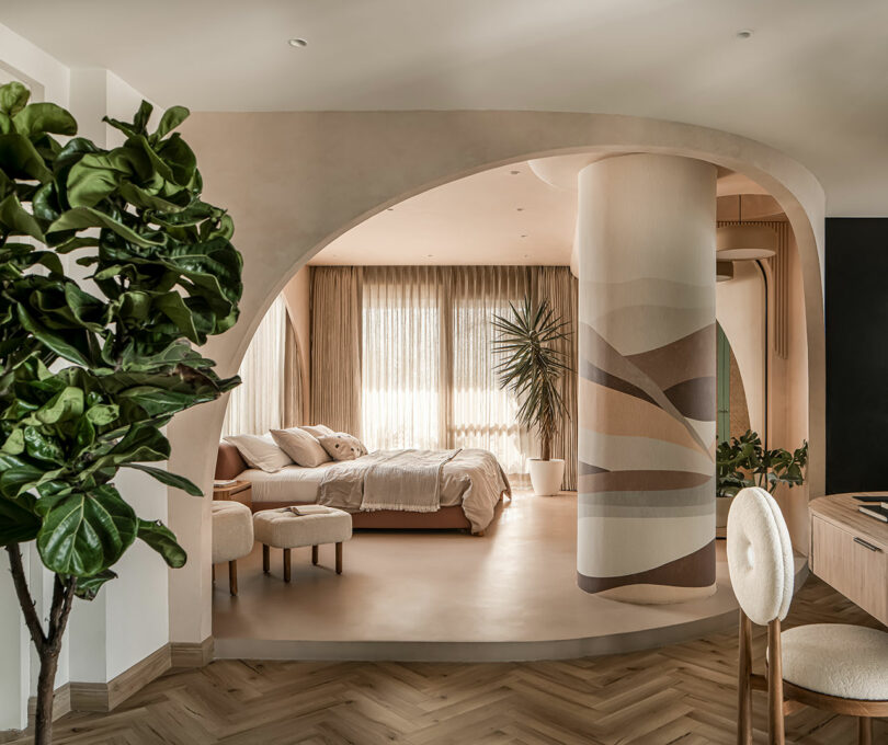 Modern bedroom with earth-toned decor, a large potted plant, and a curved architectural design. The room features a large bed, tasteful lighting, and ample natural light from floor-to-ceiling windows.