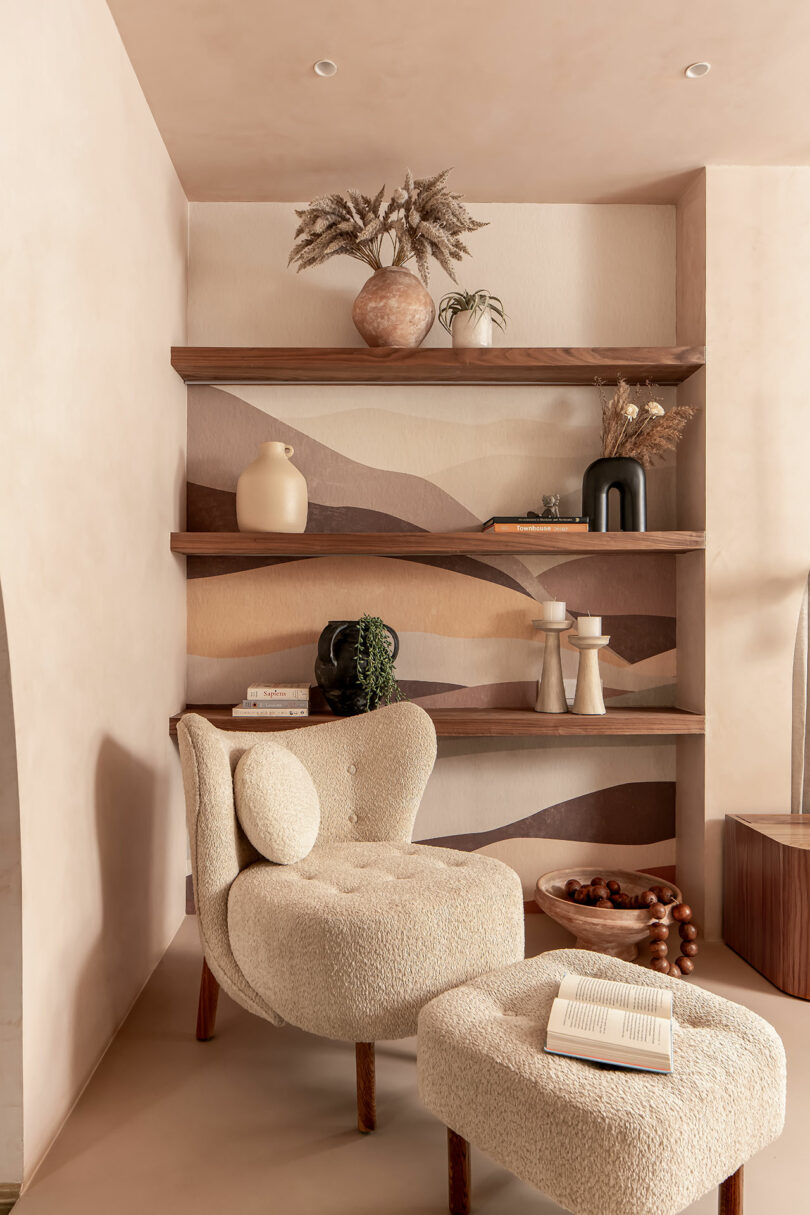 A cozy reading nook with a plush beige chair and matching ottoman, open book, wooden side table, and decorative shelves holding vases and plants against a wavy-patterned wall.