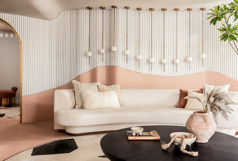 A modern living room with a white curved sofa, pink and beige decor, a black round coffee table, potted plant, and a wavy white and pink wall featuring hanging lights.