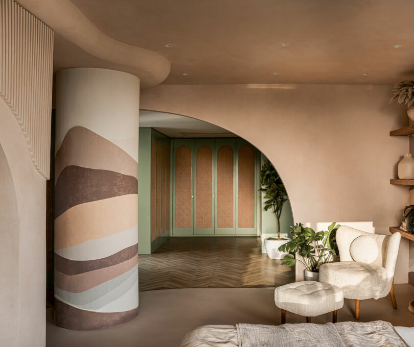 A modern room with earthy tones featuring a curved wall with a mural, wooden herringbone floor, green cabinets, plants, and beige furniture including a bed and chairs.