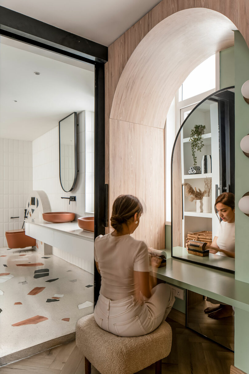 A woman sits on a cushioned stool at a vanity table with an arched mirror, in a modern, spacious bathroom featuring a double sink and contemporary decor.