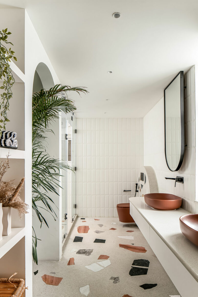 Modern bathroom with terrazzo flooring, white tiled walls, two brown vessel sinks, a large mirror, and a glass-enclosed shower. Shelving with plants and towels on the left side.