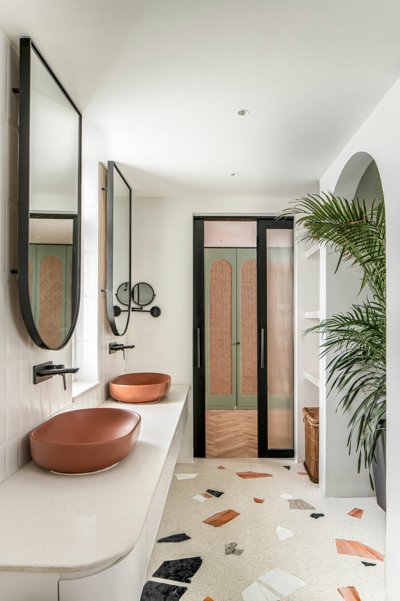 A modern bathroom features two oval mirrors, two copper sinks on a white countertop, a glass sliding door with a green and rattan design, and scattered multicolored tiles on the floor.