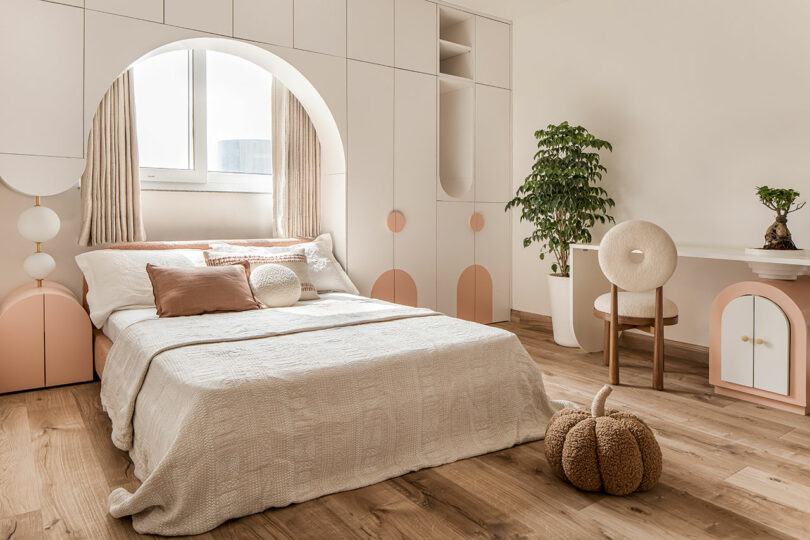 A cozy bedroom with a bed, decorative pillows, cabinets, a desk with a chair, a potted plant, a window with curtains, and a pumpkin-shaped cushion on the floor. The room has a neutral and pastel color palette.