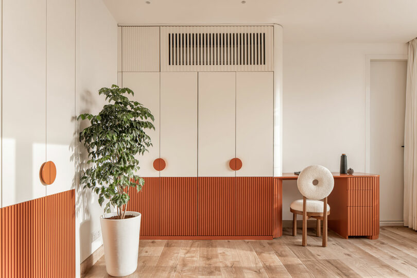 A minimalist room features white and orange cabinetry, a wooden desk with a circle chair, a potted plant, and wood flooring.