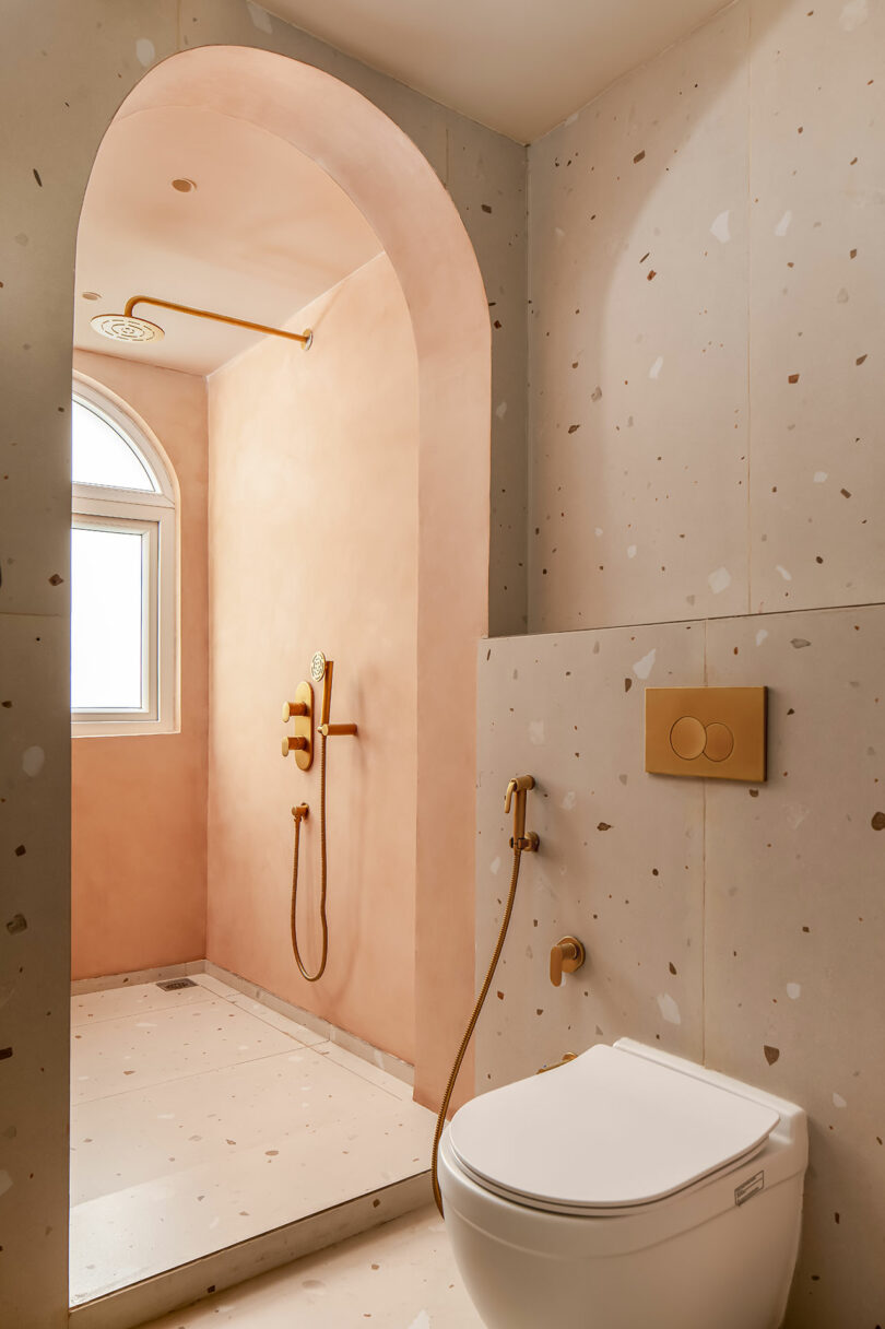 A bathroom with a toilet underneath a window, a terrazzo wall, a walk-in shower with pink walls, gold accents, and a rainfall showerhead.