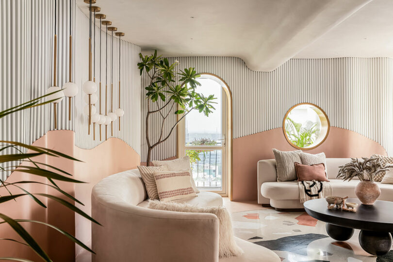 A modern living room features light-colored furniture, round mirrors, indoor plants, and a view of the outside through an arched window. The decor includes a mix of pink tones and geometric patterns.