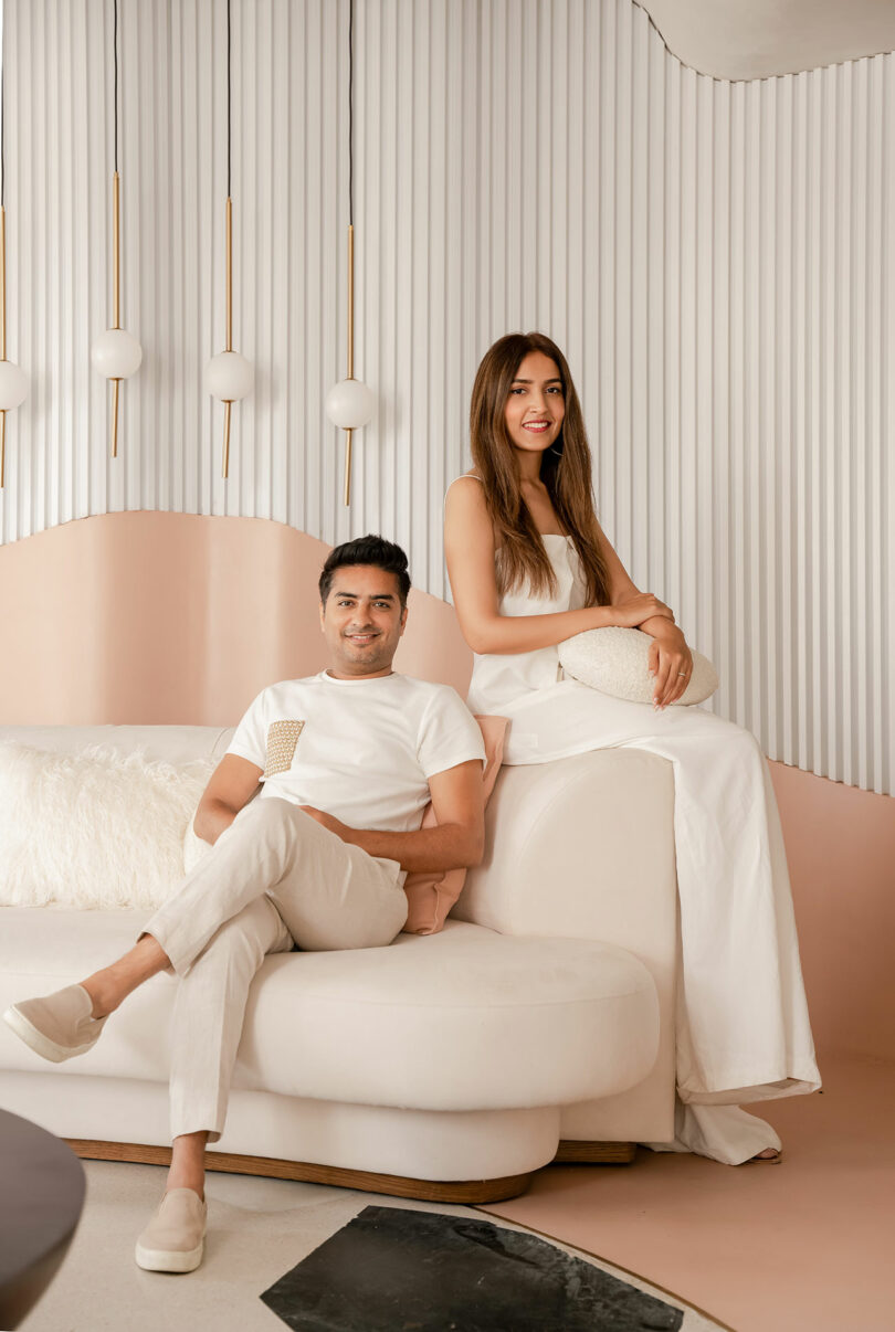 A man sits on a white couch while a woman sits on the armrest beside him. Both are dressed in white outfits, set against a background with vertical white panels and spherical hanging lights.