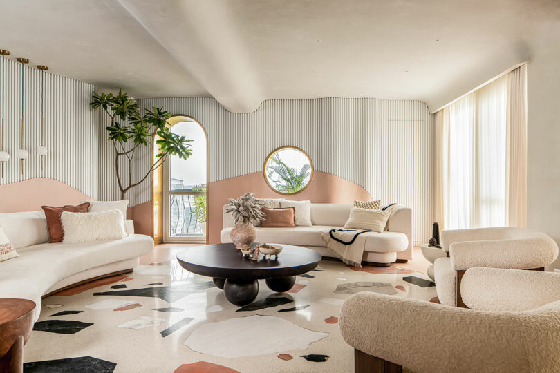 Spacious modern living room with curved white sofas, round black coffee table, abstract-patterned carpet, large potted plant, round mirror, and tall arched windows letting in natural light.