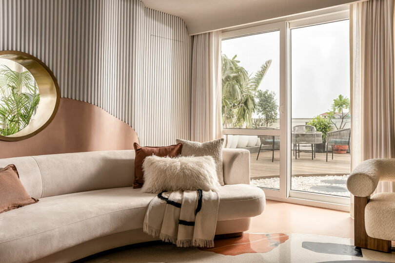 A modern living room with a curved sofa, pillows, a throw blanket, and a circular mirror. Floor-to-ceiling windows open to a patio with potted plants. Light curtains frame the window.