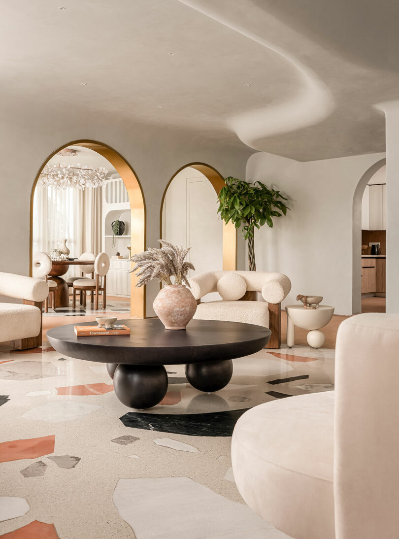 A modern living room with white curved furniture, a black round coffee table, gold archways, a terrazzo floor, and a large indoor plant. The room has a dining area visible through the archway.