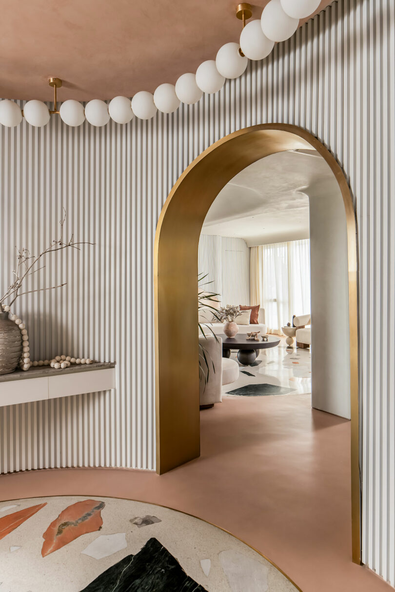 A modern interior with a gold archway, striped wall, and round ceiling light fixtures. The open doorway leads to a sitting area with plush furniture and large windows. Decorative vases sit on a shelf.