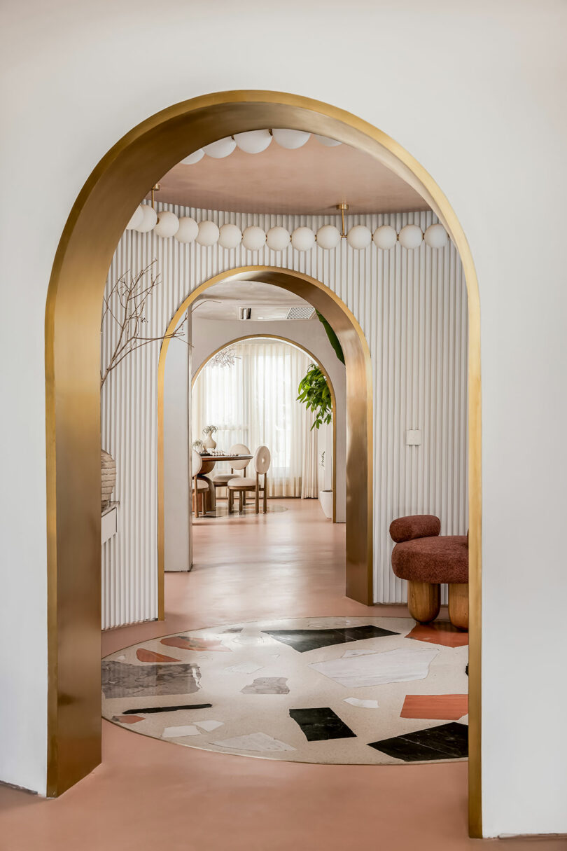 A modern interior with gold archways, abstract floor tiles, and a glimpse of a dining area with minimalist furniture and large windows in the background.