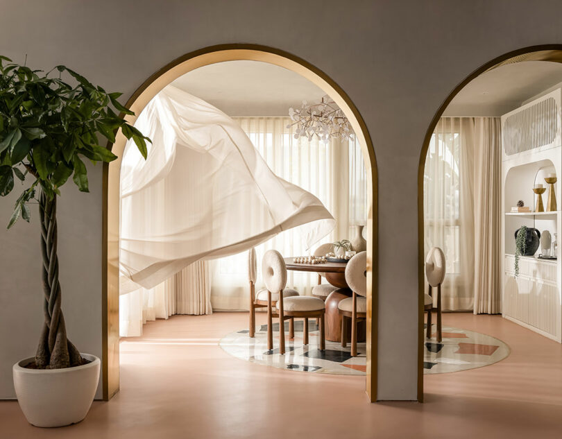 A dining room with arched doorways, beige curtains, a round table with four chairs, marble floor inset, and a potted plant in the foreground. A chandelier hangs above the table.