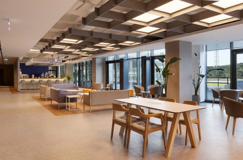 A modern office lounge area with tables, chairs, and couches under a grid-patterned ceiling. Large windows provide natural light. A counter is visible in the background.