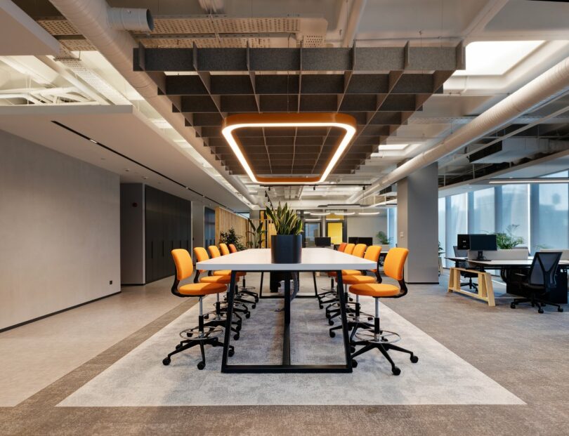 Modern conference room with a long table, orange chairs on both sides, and a central potted plant. The ceiling features a rectangular light fixture and acoustic panels. Large windows line the right side.