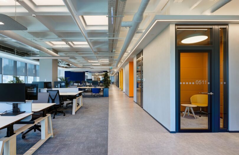 Modern open-plan office space with multiple desks, computer monitors, and a hallway leading to private meeting rooms. Bright overhead lighting and large windows provide abundant natural light.