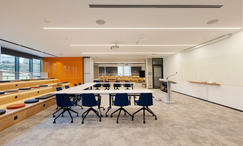 A modern classroom with tiered wooden seating, blue swivel chairs around a rectangular table, a podium, front projection screen, and a wall-sized whiteboard. Large windows allow natural light in.