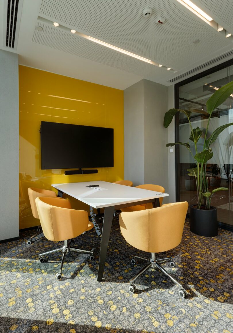 A small modern conference room with a white table, yellow chairs, a mounted TV, large potted plants, and a patterned carpet.