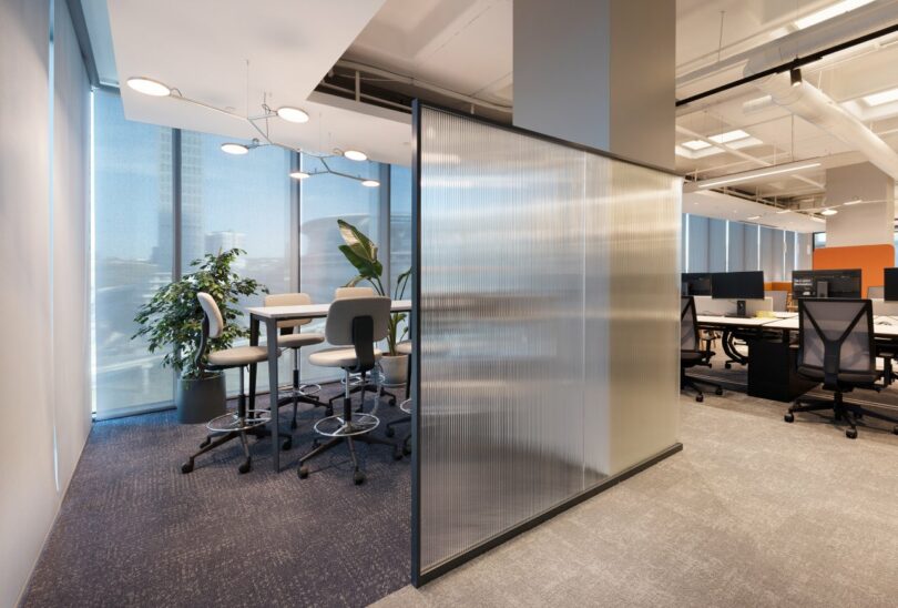 Modern office space with a high table, stools, and a large plant on the left; partition separates this area from an open-plan workspace with desks and computers on the right.