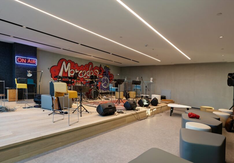 A modern music studio with instruments, microphones, and amplifiers on a stage. A neon "On Air" sign is mounted on the wall next to a colorful graffiti mural. Minimalist seating is arranged in front.