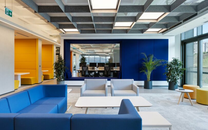 Modern office lounge area with blue and grey seating, white tables, large windows, and potted plants; an adjacent meeting room is visible through a glass partition.