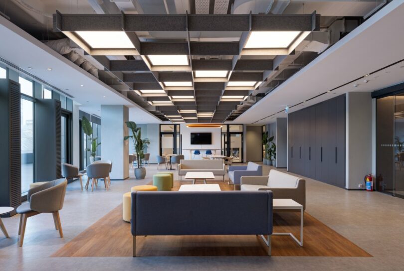 A modern, spacious office lounge area with various seating options, a large overhead lighting fixture, and a television on the far wall.