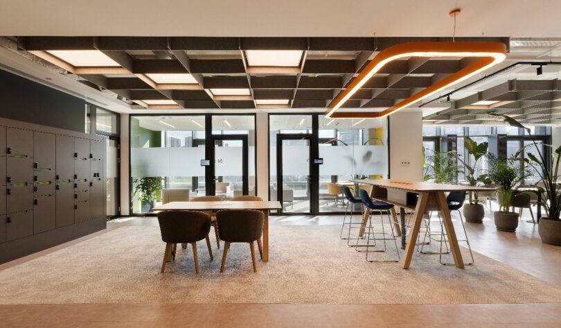 A modern office lounge area featuring a long table with high chairs, a smaller table with four chairs, lockers, and large floor-to-ceiling windows allowing natural light to enter.