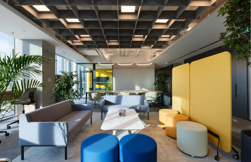 A modern office lobby features colorful furniture including sofas, stools, and partition screens. The space is well-lit with natural light and ceiling lights, and has several potted plants and large windows.