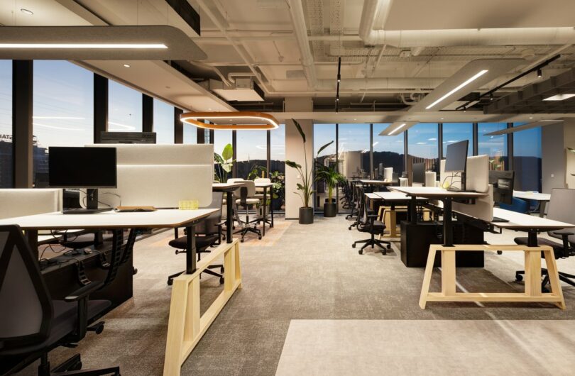 A modern office space with open workstations, large windows, multiple computer monitors, chairs, and indoor plants, illuminated by overhead lighting.