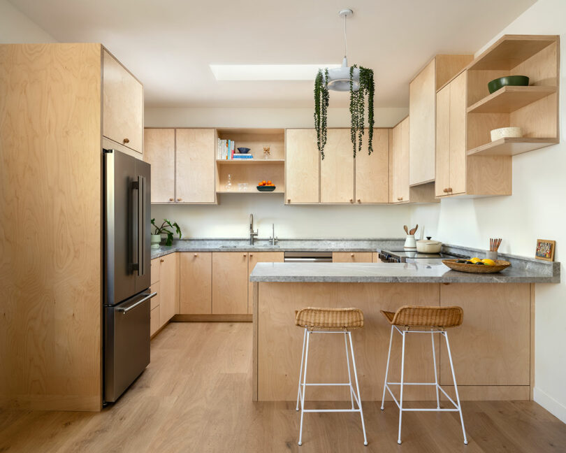 Modern kitchen with light wood cabinets, grey countertops, and stainless steel appliances. Two wicker stools are placed at the island, and hanging plants add a touch of greenery.