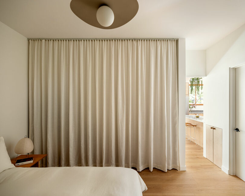 A minimalist bedroom featuring a bed with a white duvet, a small nightstand with a lamp, and a large beige curtain separating the room from an adjacent sink area. The floor is wooden.