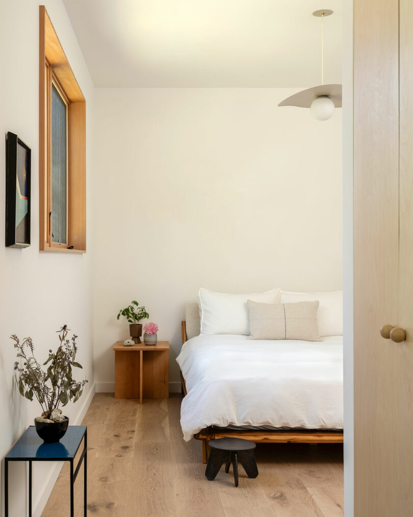 A minimalist bedroom with a white bed, light wood floors, a side table with a plant and a vase, and a modern light fixture. A small painting hangs on the wall near a window with a wooden frame.