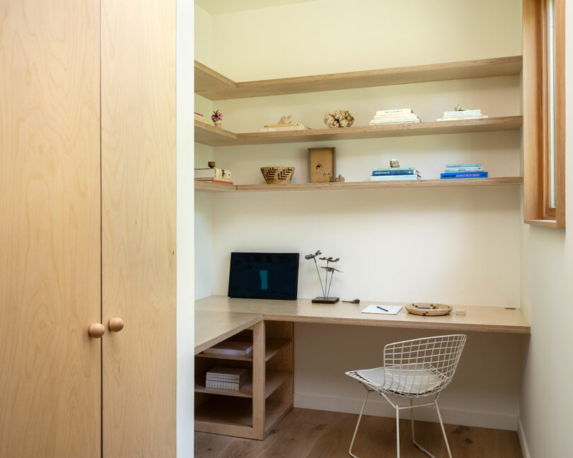 A small home office with a built-in wooden desk, open shelves holding various items, and a white wire chair. A laptop, notebook, and decorative objects are on the desk. Light wooden flooring.