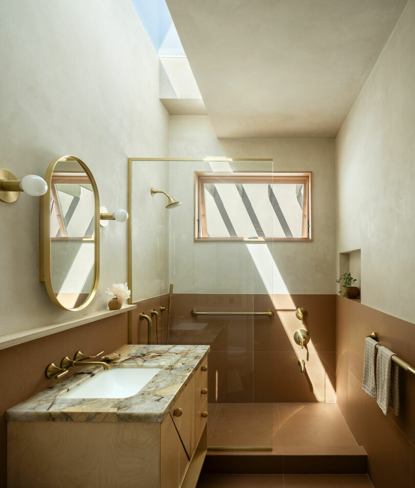 A modern bathroom with natural light, featuring a marble-top vanity, gold fixtures, an oval mirror, a glass-enclosed shower, and a skylight overhead.