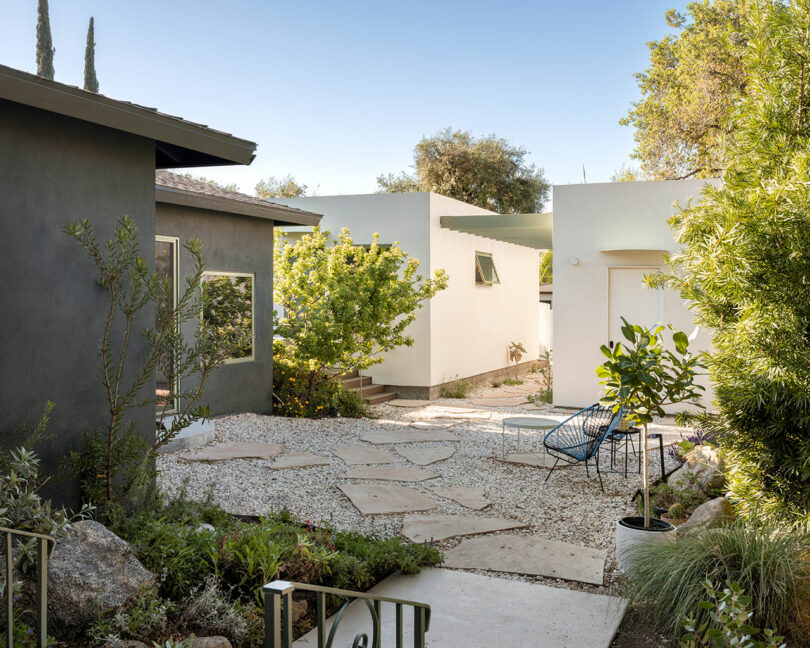 A modern outdoor space featuring two minimalist buildings, a stone pathway, and greenery including trees and shrubs. A blue chair and a small table are situated on the gravel area.