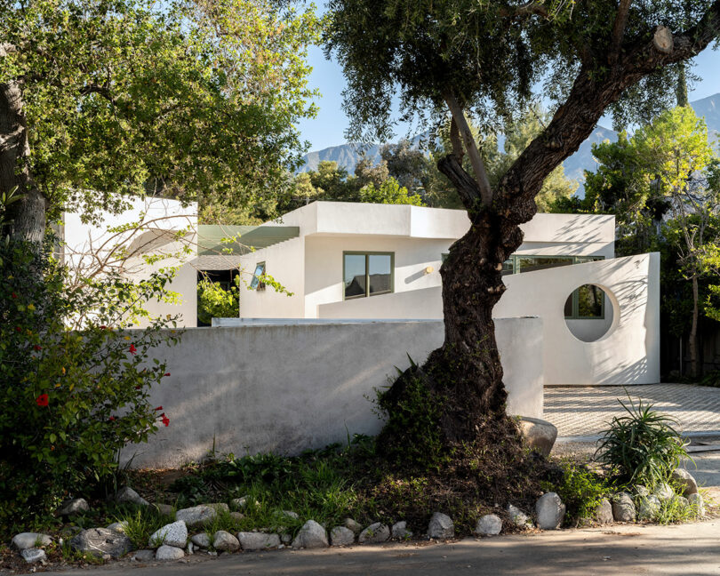 Modern white house with flat roofs, large windows, and a circular cut-out detail, surrounded by trees and greenery, with mountains visible in the background.