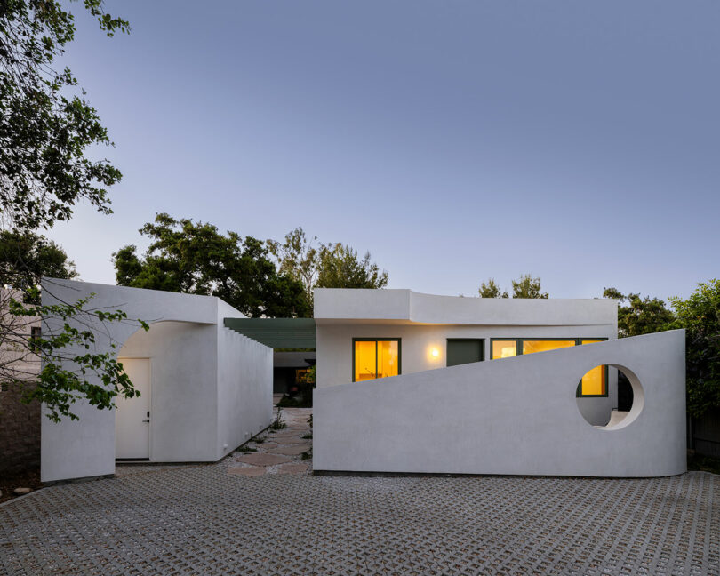 Modern white house with geometric design features, including a large circular cutout in the exterior wall. The façade is minimalistic, and the house is surrounded by trees. The driveway is paved with tiles.