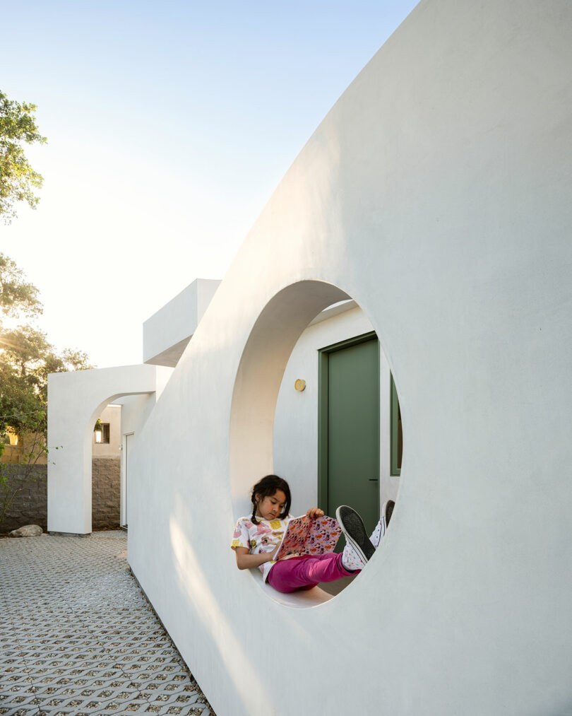 A person sits in a circular opening of a white wall, reading a book in a modern architectural setting with a green door in the background.