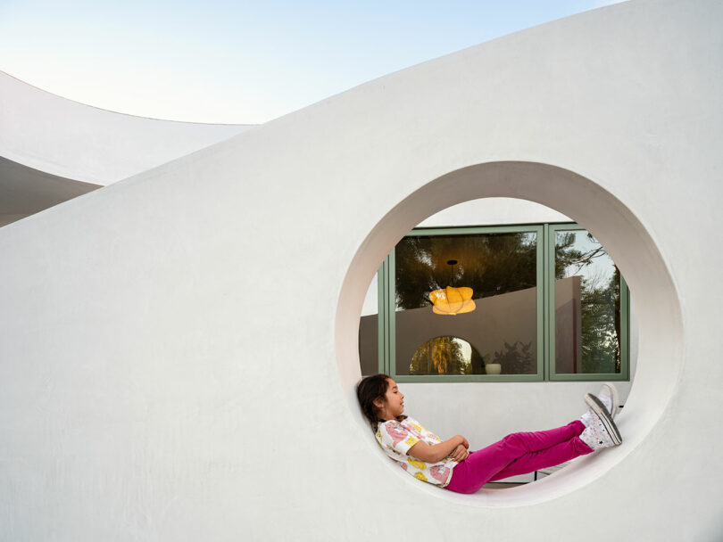 A child in pink pants and a white shirt reclines inside a circular opening of a white modern building with green-framed windows.