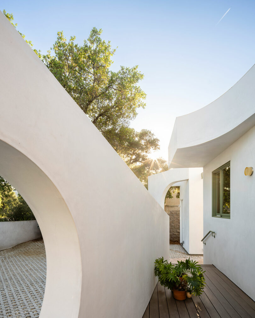 A modern white building with curved walls, an arch, and a deck, surrounded by trees and a clear sky.