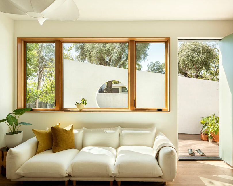 A sunlit living room features a white sofa with yellow pillows, a wooden coffee table with plants, large windows, and an open door leading to a wooden deck with shoes placed near the entrance.