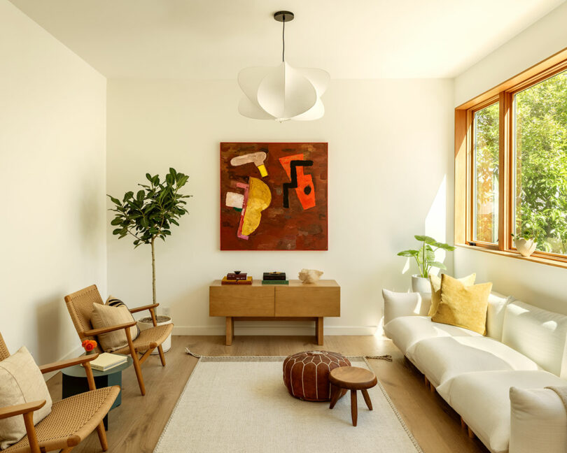 A bright living room with a large window, modern furniture including a white sofa, wooden chairs, a low cabinet, and abstract art on the wall. A potted plant stands in the corner, and a pouf and small stool are on the rug.