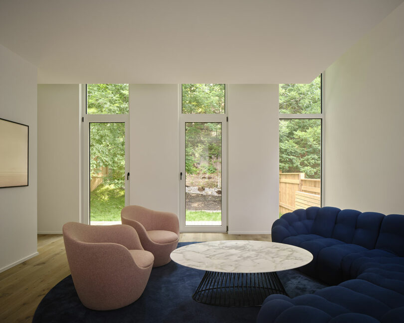 A modern living room features large windows, a blue sectional sofa, two pink armchairs, and a round marble coffee table on a navy rug, with a view of a grassy backyard.