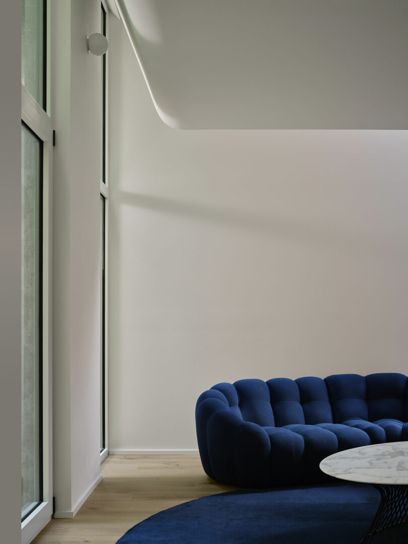 Minimalist interior featuring a navy blue tufted sofa, large windows, and a white marble coffee table on a round blue rug.