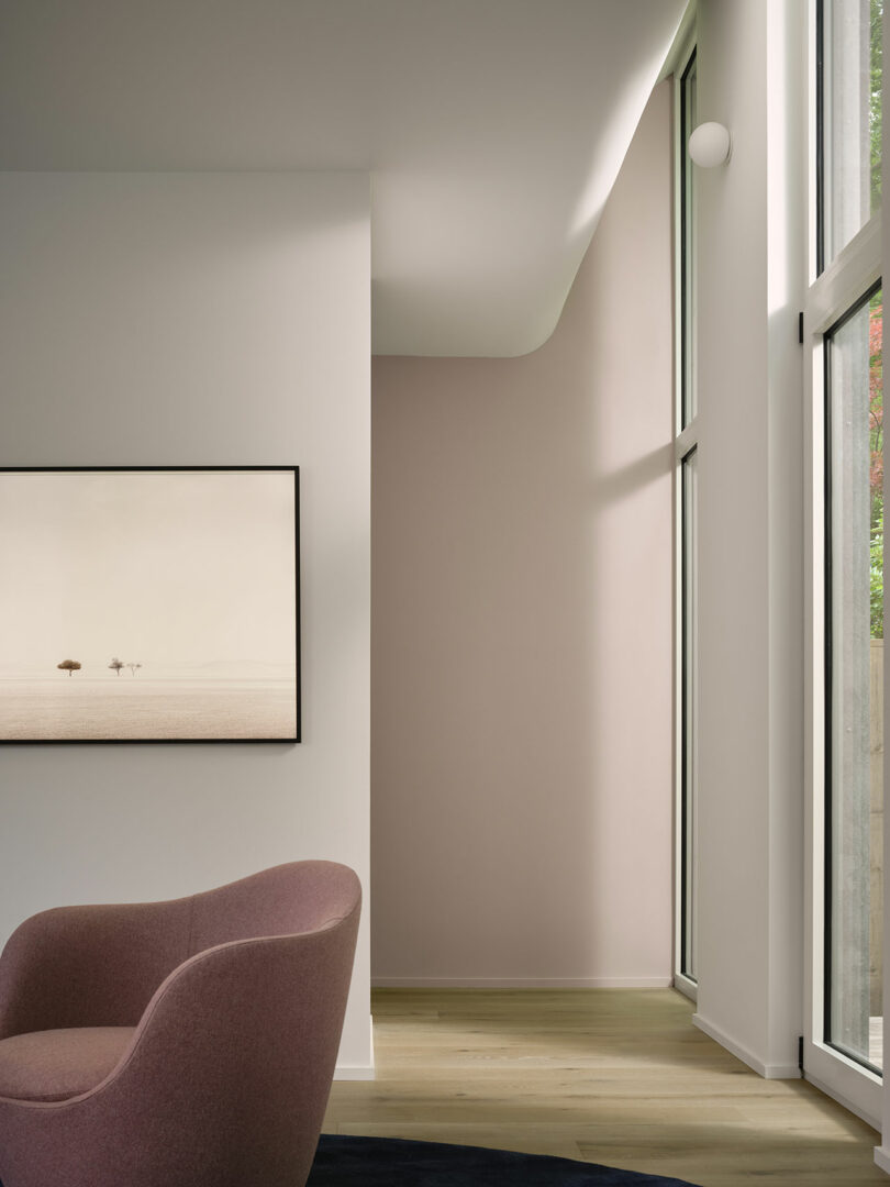 Modern interior with large windows, a pink armchair, wooden flooring, and a framed minimalist painting on a white wall.