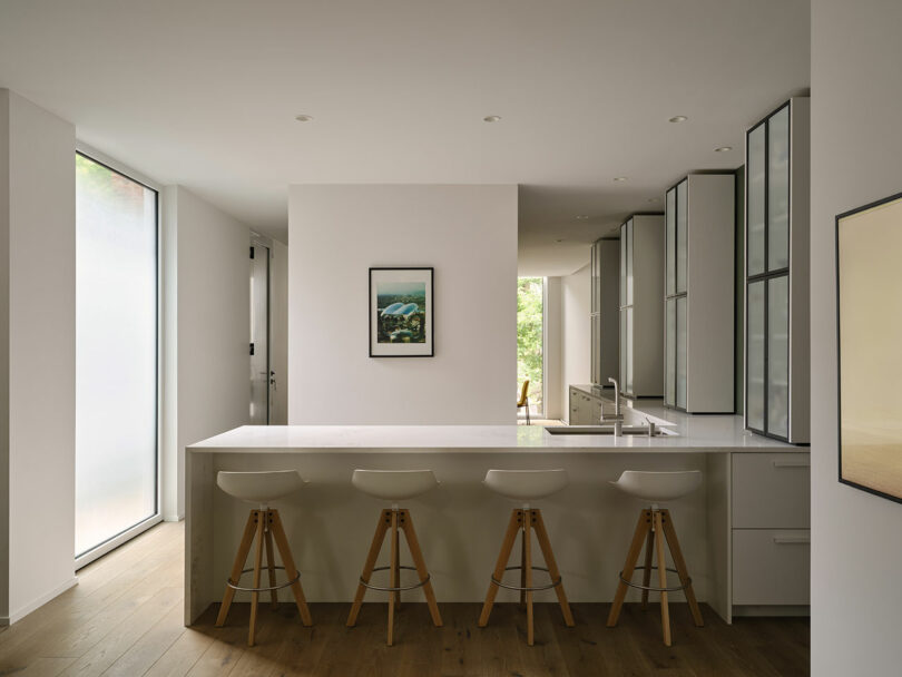 Modern kitchen with a long white countertop, four wooden barstools, minimalist decor, large windows, and sleek cabinets. Framed art hangs on the wall above the counter.