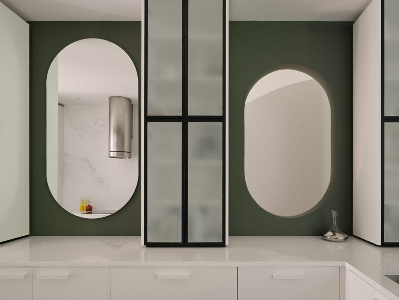 A contemporary kitchen with white cabinets and green walls features two oval mirrors and a stainless steel vent hood. A small vase is placed on the countertop on the right side.