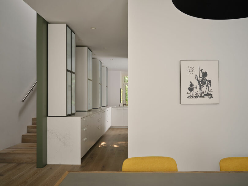 Modern kitchen interior with white cabinets, a green accent wall, wooden flooring, and a staircase with a handrail. A framed black-and-white artwork is hung on the adjacent white wall.