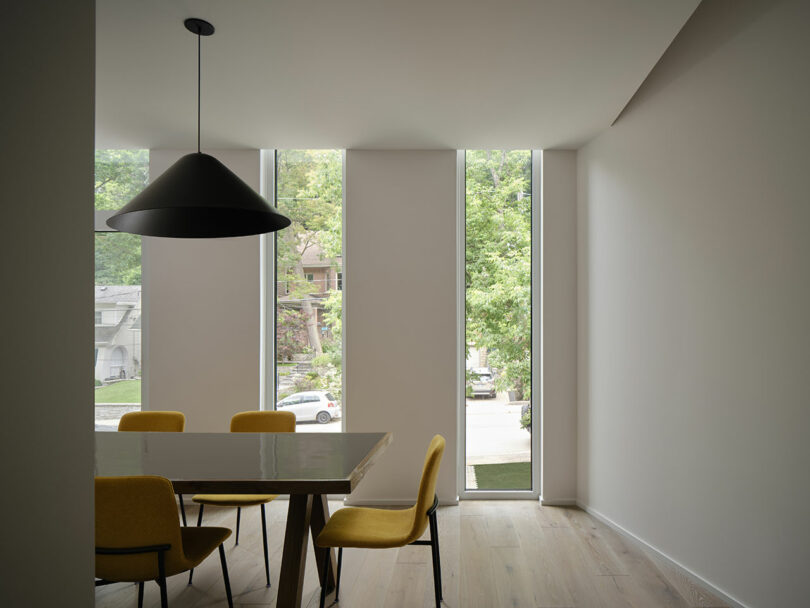 A modern dining room with a glass table, yellow chairs, large vertical windows, and a black pendant light fixture.